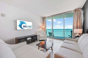 Une chambre dâhĂŽtel moderne dotĂ©e dâun grand balcon, dâun canapĂ©, dâune table basse et dâun tĂ©lĂ©viseur affichant le logo de Big Fish Rentals.
