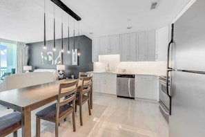 A modern kitchen with white cabinets, stainless steel appliances, a dining area with a wooden table and chairs, and pendant lighting.