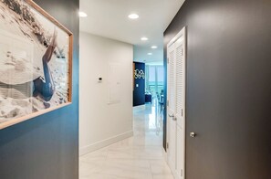 A hallway with a framed artwork on the left, a door with louvered shutters on the right, and a dining area in the background.
