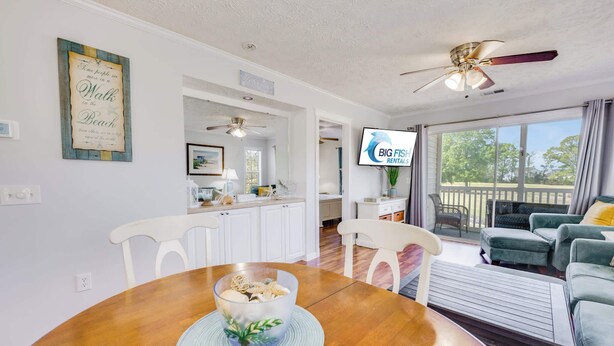 A dining area with a wooden table, chairs, and a bowl of snacks. There is a kitchen area with white cabinets and a ceiling fan. A TV is mounted on the wall, and a balcony with outdoor furniture is visible. - NEW! Large 2BR| River Oaks Golf | Pools | Hot Tubs (Myrtle Beach)