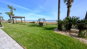 Une pelouse bien entretenue, agrémentée d’une balançoire et d’une pergola, de palmiers et offrant une vue sur la plage.
