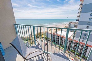 A balcony with a view of the beach and ocean, a parking area, and a multi-story building with balconies.