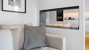 Un saló modern amb un sofà, una llar de foc i una cuina visible a través d'una separació de vidre.