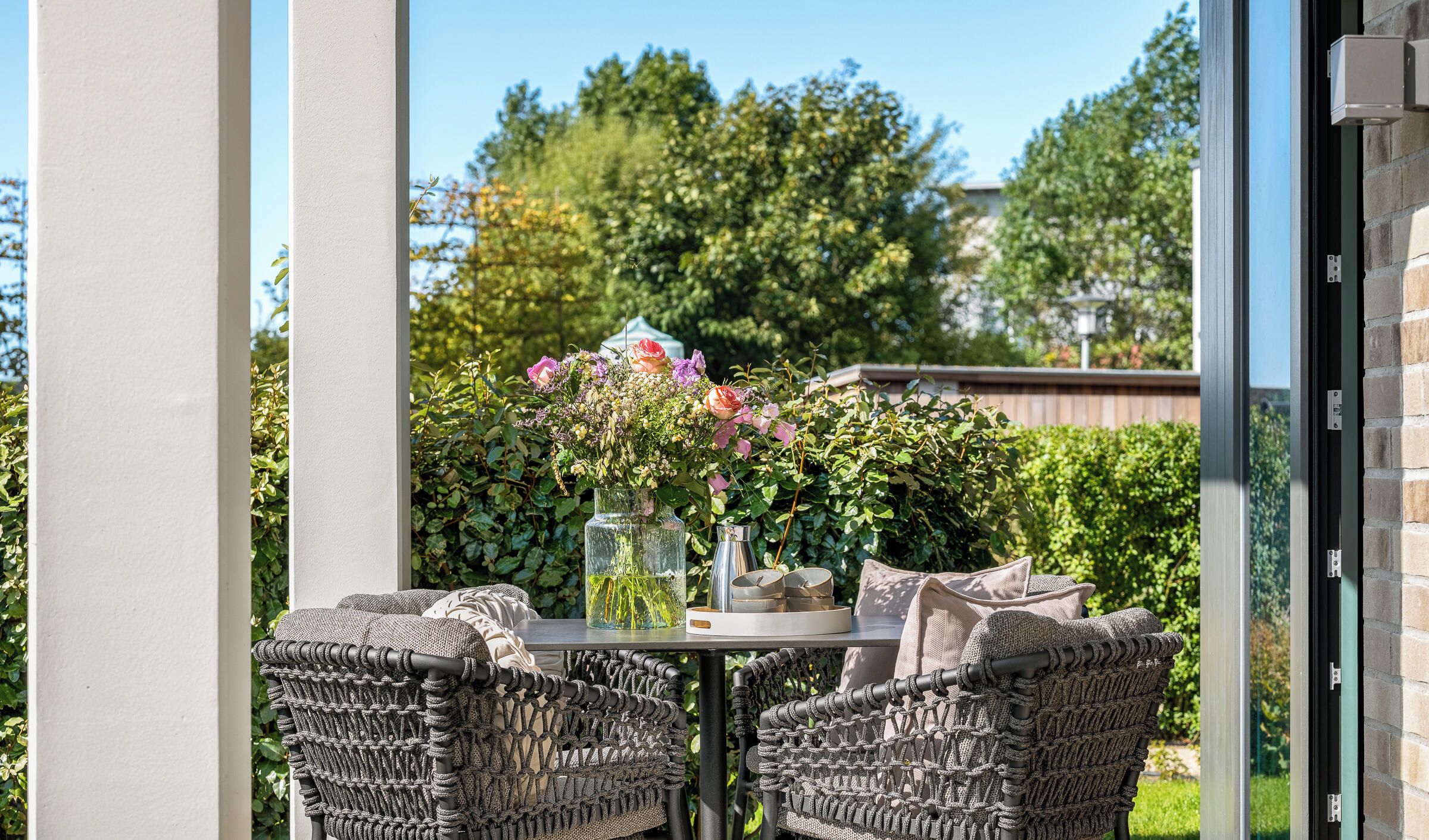 Ein Sitzbereich im Freien mit Korbstühlen und einem Tisch, einer Vase mit Blumen sowie Blick auf einen Garten und Bäume.