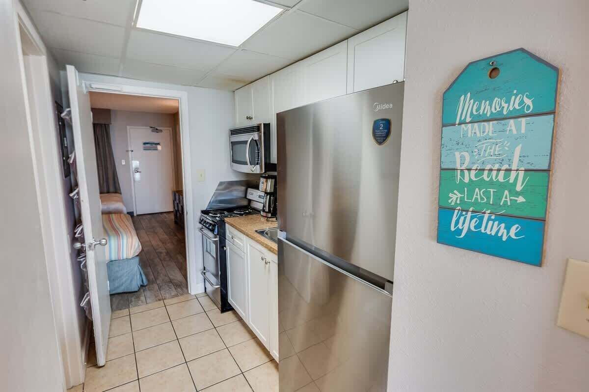A compact kitchen with a stainless steel refrigerator, microwave, and stove. A teal sign on the wall reads "Memories Made at the Beach Last a Lifetime."