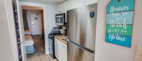 A compact kitchen with a stainless steel refrigerator, microwave, and stove. A teal sign on the wall reads "Memories Made at the Beach Last a Lifetime."