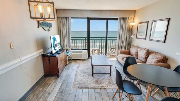 A room with a wooden floor, a sofa, a table with chairs, a TV, and a view of the beach through large sliding doors. - New 1BR Direct Oceanfront  Sandcastle South, Pools (Myrtle Beach)