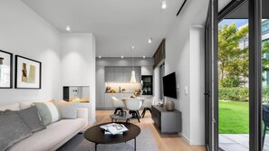 A modern living room with a sofa, coffee table, and a dining area with chairs. There is a kitchen in the background with a TV and a window showing a garden.