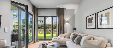 A modern living room with a sofa, coffee table, and a view of a garden through large glass doors.