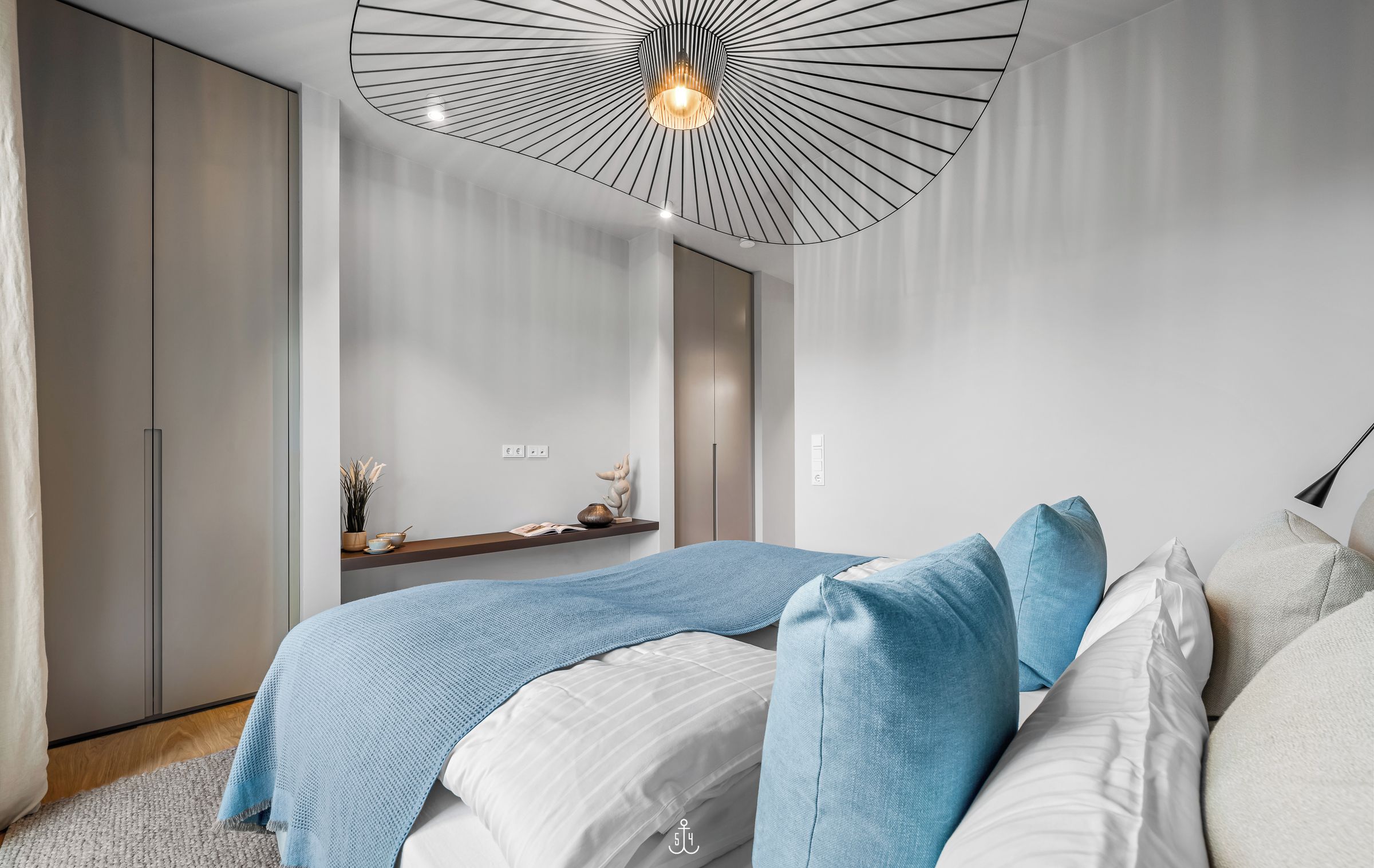 Un dormitorio moderno con cama, almohadas azules y blancas, una lámpara colgante y una pequeña repisa con adornos.