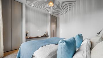 Un dormitorio moderno con cama, almohadas azules y blancas, una lámpara colgante y una pequeña repisa con adornos.