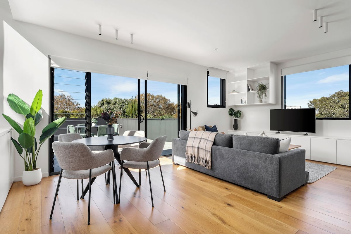 A modern living room with a grey sofa, a round dining table with chairs, a flat-screen TV, and large windows offering a view of the outdoors.
