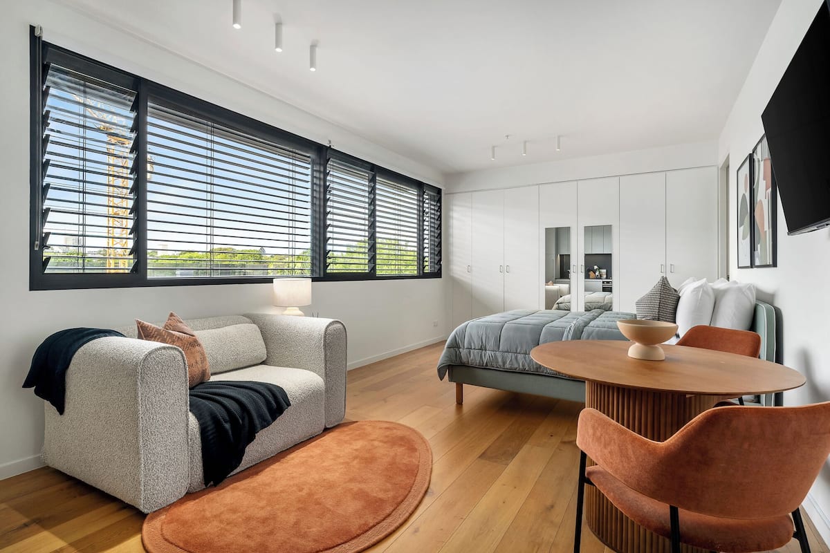 A modern bedroom with a large window, a bed, a sofa, and a dining area.