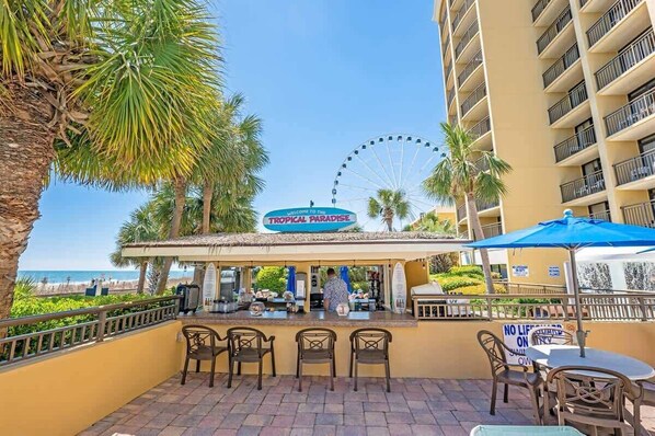 A Tropical Paradise bar with outdoor seating, a Ferris wheel in the background, and a view of the beach. - Direct Ocean Front, The Pavilion, Pools, Boardwalk (Myrtle Beach)