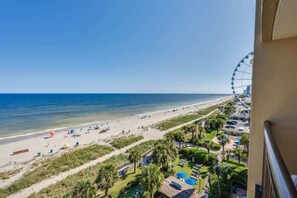 A beach view with a Ferris wheel, beach umbrellas, and a resort area with buildings and palm trees. - Direct Ocean Front, The Pavilion, Pools, Boardwalk (Myrtle Beach)