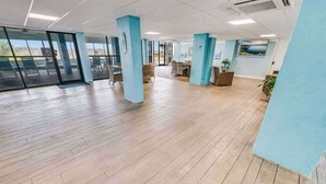 Un espacio interior amplio con suelo de madera, pilares azules y una zona de estar con sillas y mesas.