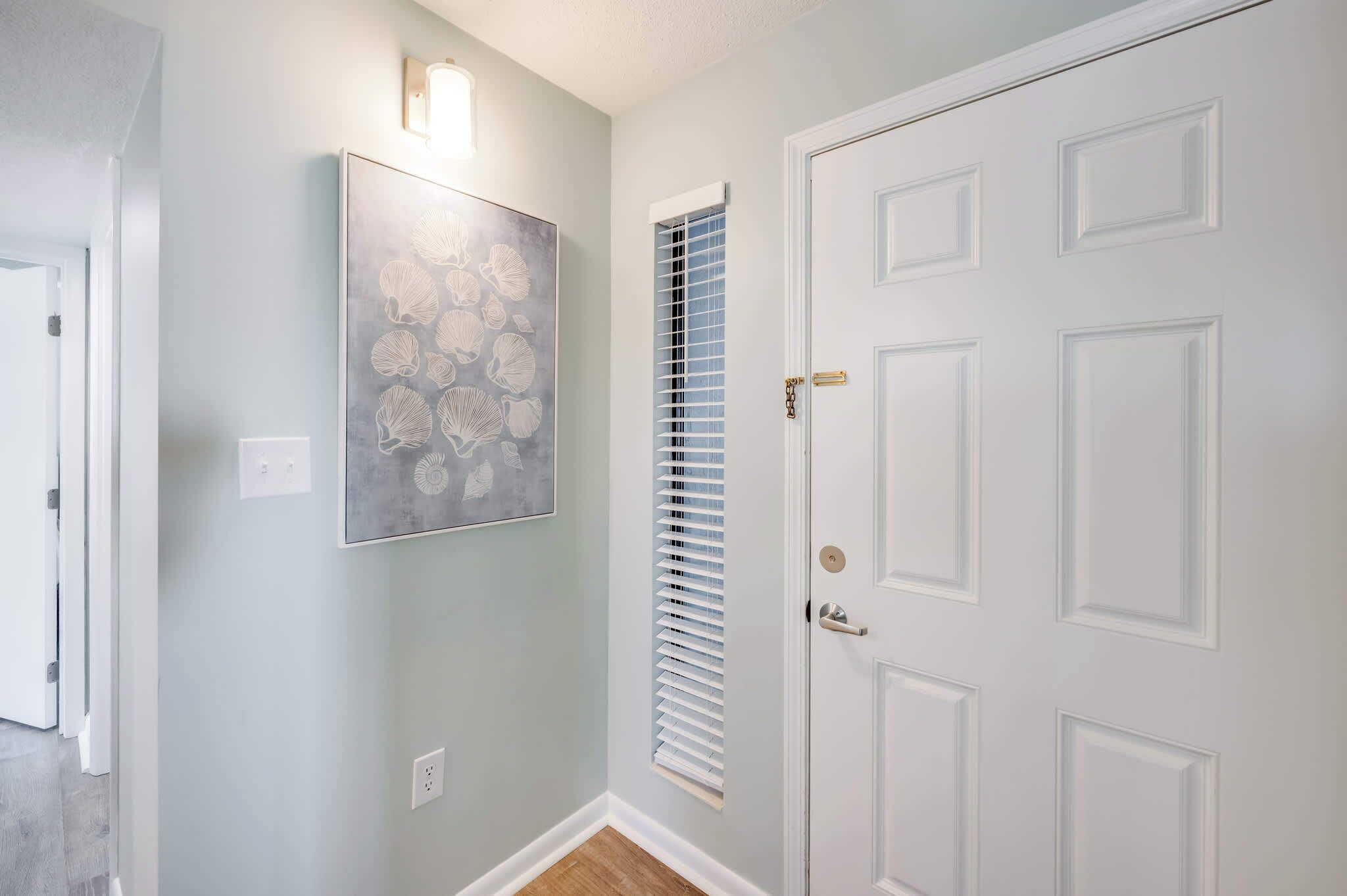 A hallway with a light blue wall, a white door with a gold handle, and a window with blinds.