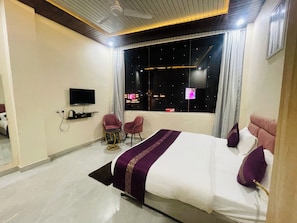 A modern hotel room with a large bed, a TV, a small table, and a window with a view of the city at night. - The Kashi Stays (Varanasi)