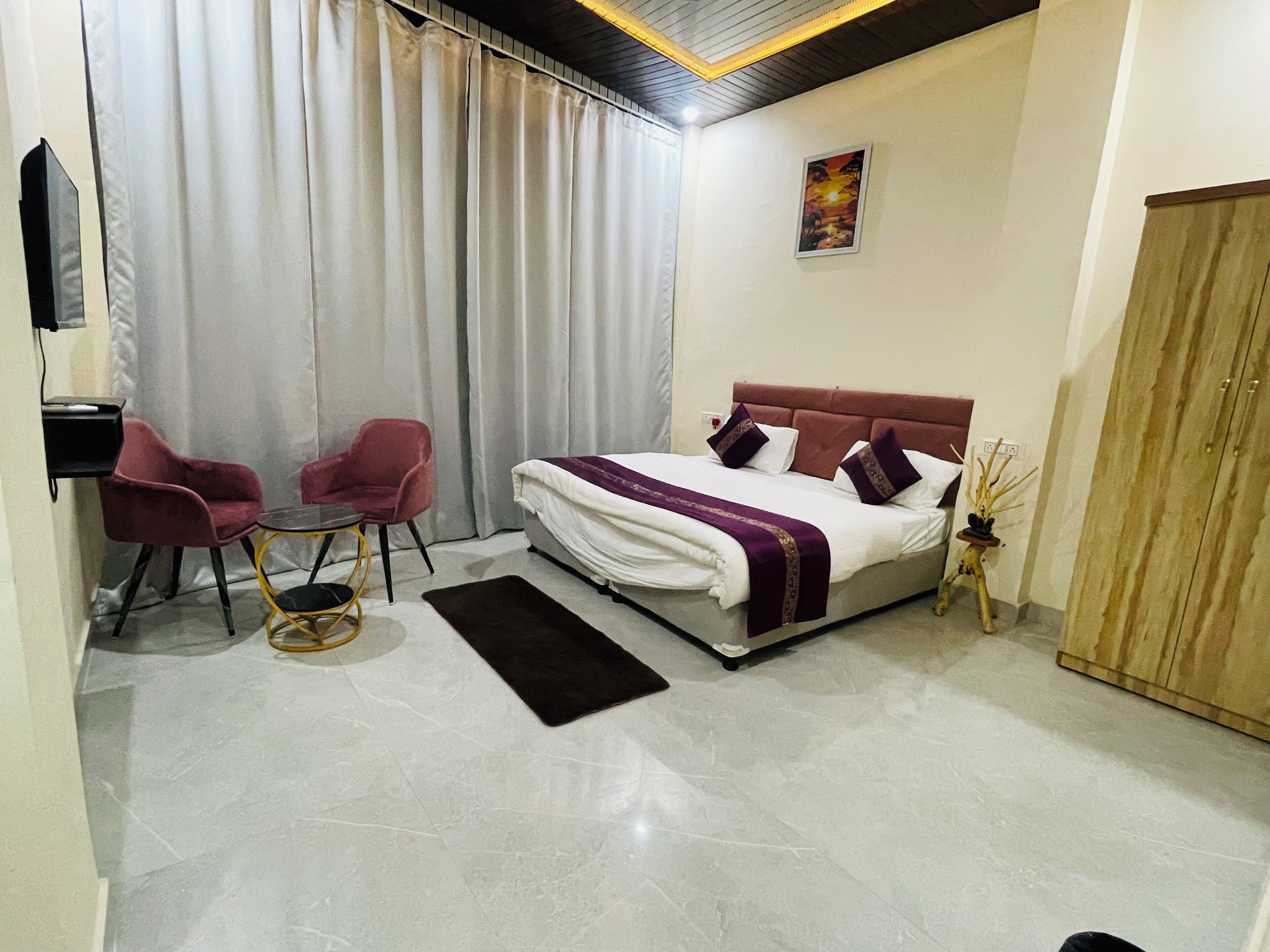 A hotel room with a bed, two chairs, a small table, a rug, a wooden wardrobe, and a painting on the wall.