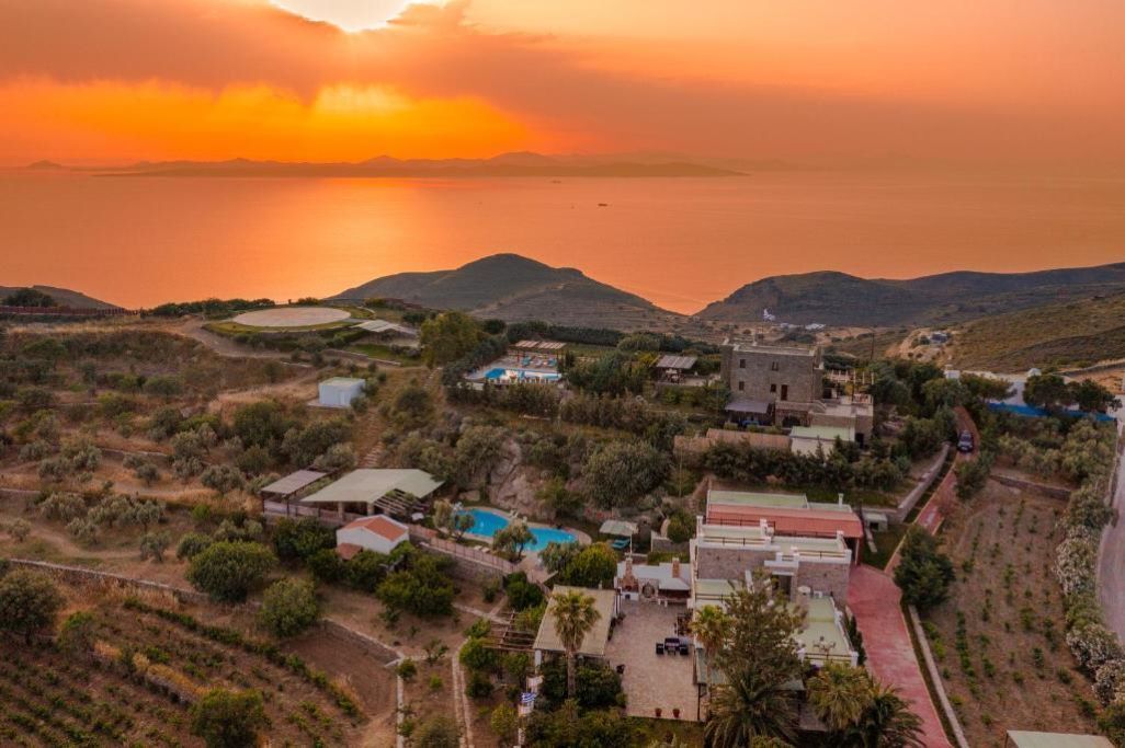 An aerial view of a coastal village with buildings, a swimming pool, and a view of the sea and hills.