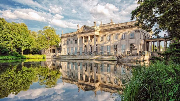 Een statig gebouw met klassieke architectuur, een reflecterend waterlichaam ervoor en weelderige groene beplanting eromheen.