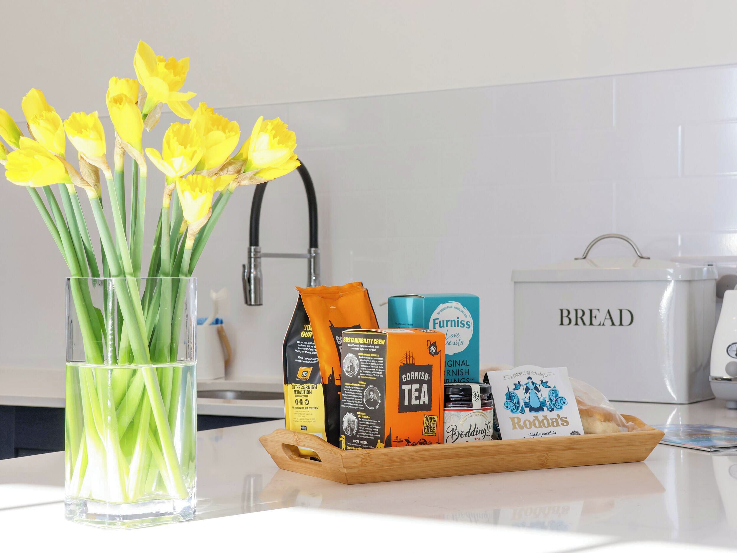 A kitchen counter with a glass vase of yellow flowers, a wooden tray with various food items, and a white bread container.