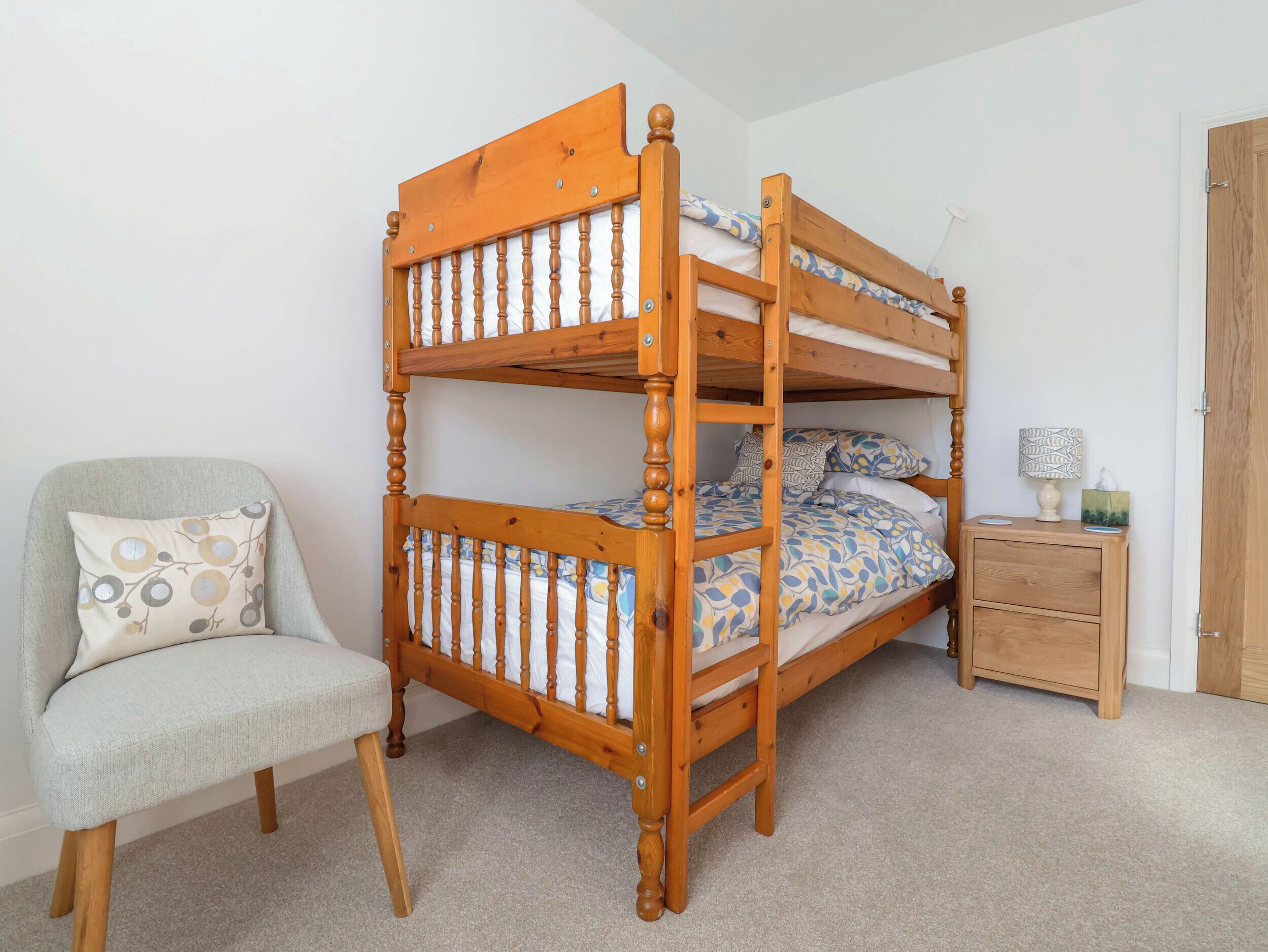 Una cama litera con estructura de madera, una silla con cojín, una mesita de noche con lámpara y una puerta al fondo.