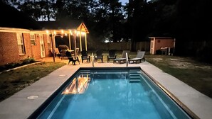 A rectangular swimming pool with a wooden deck and steps leading into it. There are lounge chairs and a covered patio area with string lights.