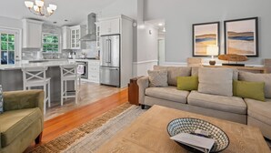A modern kitchen with white cabinets, a stainless steel refrigerator, and a bar area. There is a beige sofa with green cushions in the living area, and a wooden coffee table.