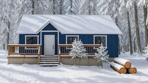 Una cabaña azul con una puerta blanca, porche de madera y escalones que conducen a la entrada, rodeada de árboles y troncos cubiertos de nieve.