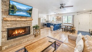 Una sala de estar moderna con chimenea, un televisor montado en una pared de piedra, una cocina al fondo y un ventilador de techo.