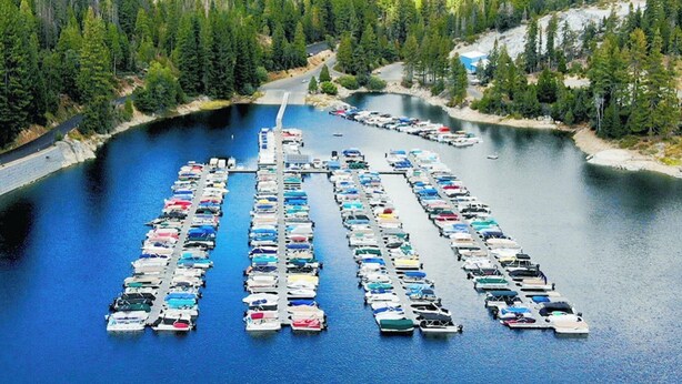 Una marina con numerosos barcos amarrados en un lago tranquilo rodeado de un bosque denso y montañas.