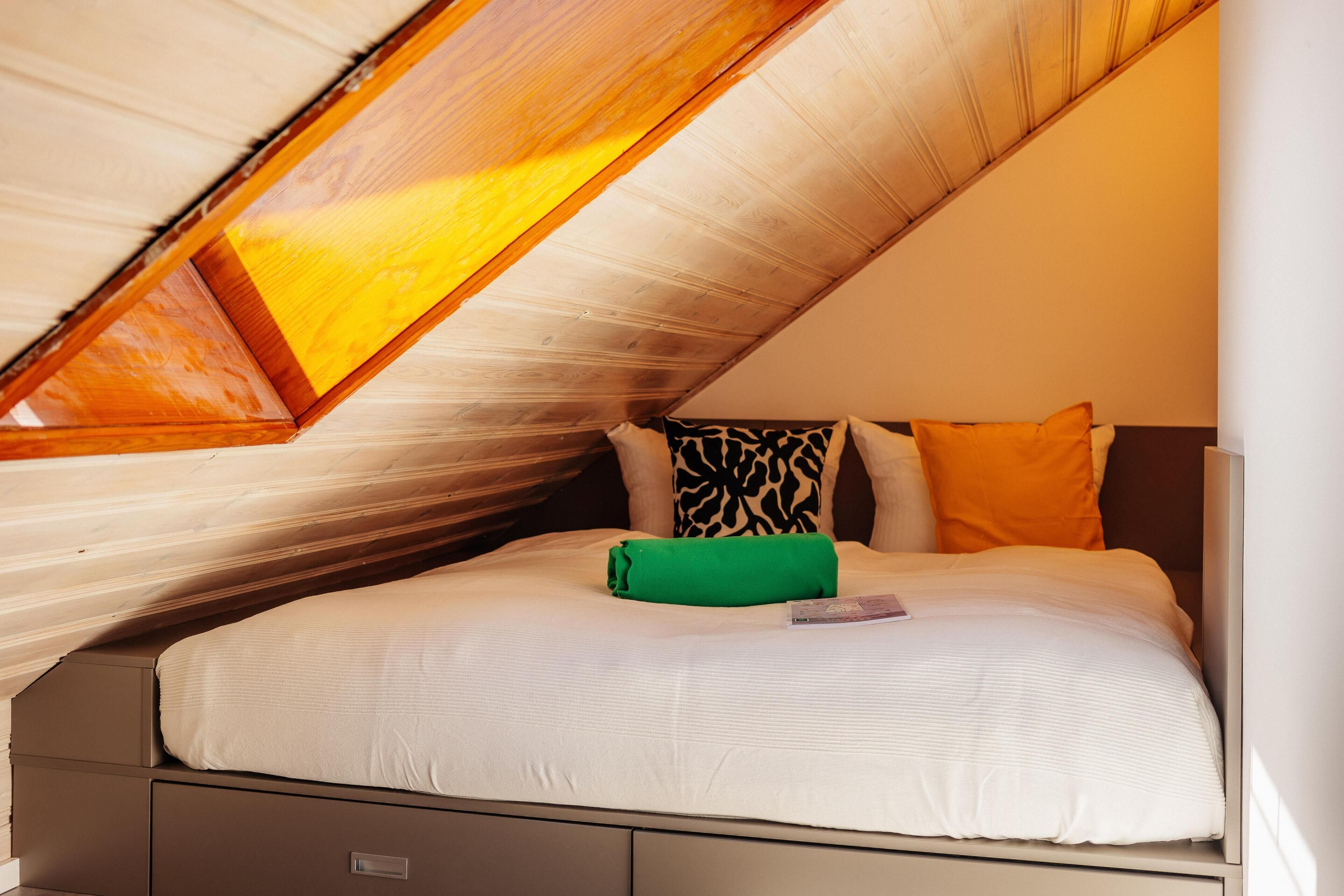 Ein Schlafzimmer mit schrägverdeckter Decke, einem Bett mit weißer Bettwäsche, einem grünen Kopfkissen und einem gemusterten Kissen.