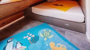 Une chambre avec un plafond en pente, un lit recouvert d’une couverture jaune, un tapis bleu orné de motifs d’animaux marins et une lampe rouge en forme de fleur.