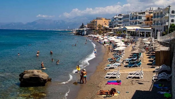 A beach with sun loungers and umbrellas, a rocky shoreline, and buildings in the background. - Chersonissos Central Apartment (Hersonissos)