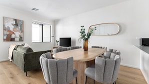 A modern dining area with a round table, grey upholstered chairs, a vase with plants, a large mirror, and a flat-screen TV. - Stafford St Apartments by Urban Rest (Adelaide)