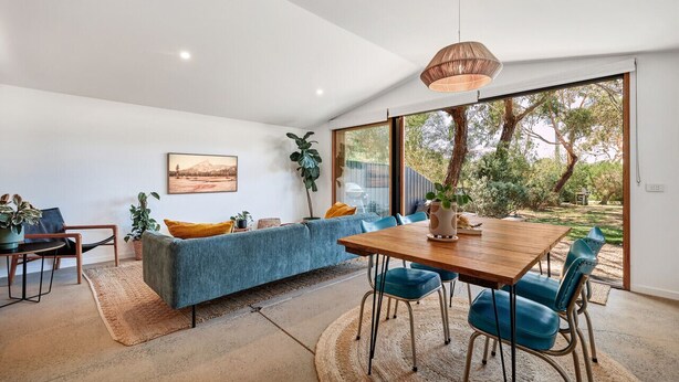 A modern living room with a wooden dining table, blue chairs, a blue sofa, and a large window offering a view of greenery. - Secluded Bellbrae sanctuary (Bellbrae)