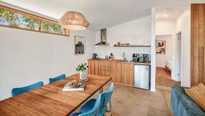 A modern kitchen with wooden cabinets, a dining area with a wooden table and blue chairs, and a large window with a view of trees. - Secluded Bellbrae sanctuary (Bellbrae)