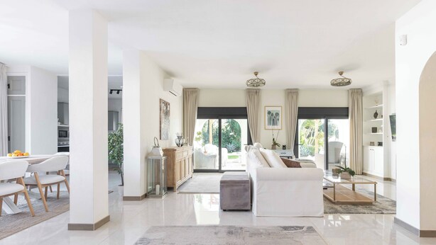 A spacious living area with a white sofa, a coffee table, and a dining table with chairs. There is a large window with curtains, a chandelier, and a view of greenery outside. - Villa Hope -Luxury 3-bedroom villa with AC, WiFi in lovely Buenas Noches (Buenas Noches)