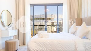Ein Hotelzimmer mit einem großen Bett, einem runden Spiegel, einem kleinen Hocker und Blick auf die Stadt durch große Fenster.