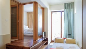 A hotel room with a wooden wardrobe, a bed with white bedding, a television on a wooden stand, and a view of the outdoors through a glass door.