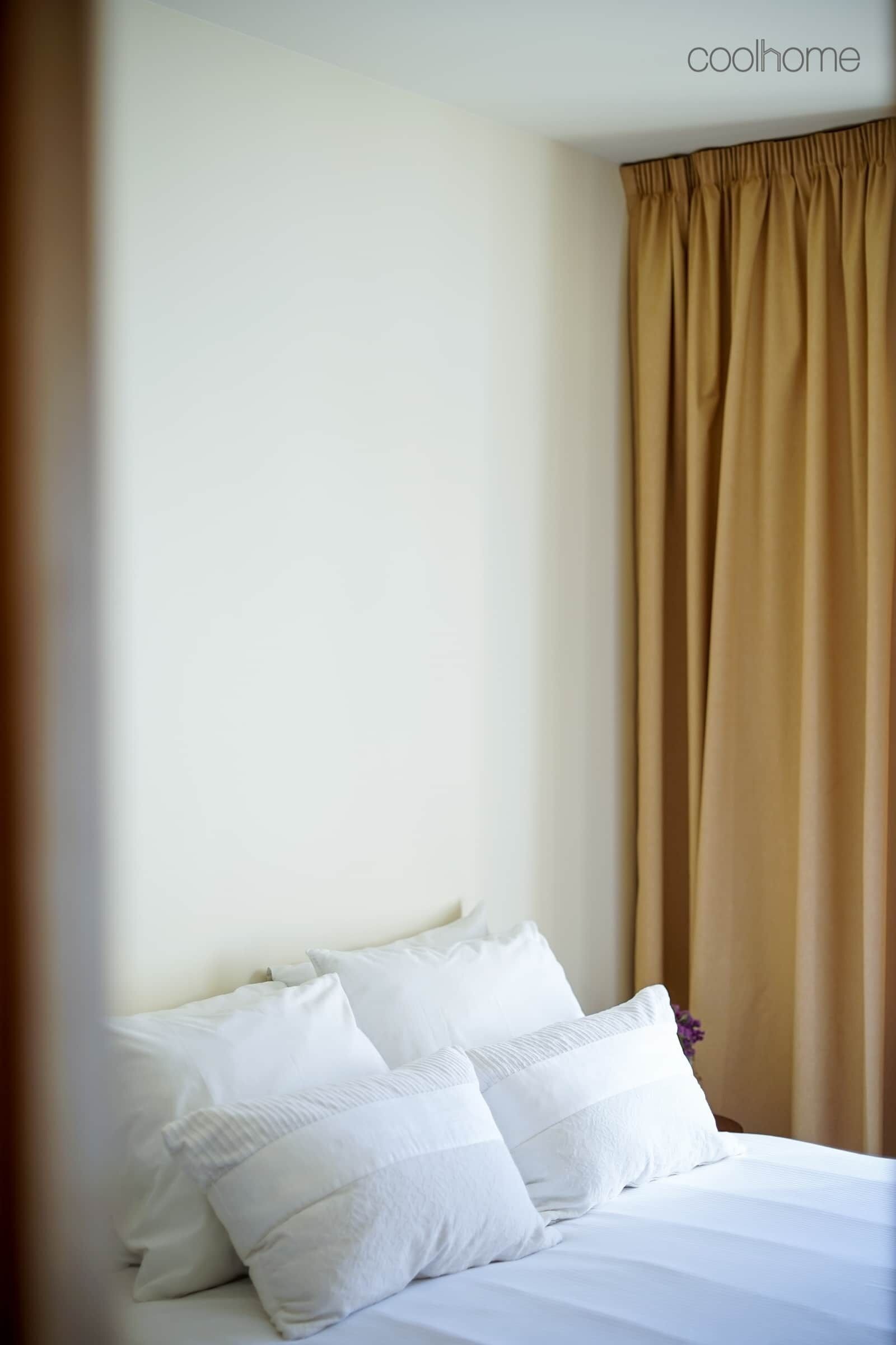 A neatly made bed with white linens and pillows, a beige curtain, and a plain wall.