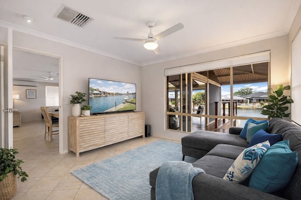 A modern living room with a grey sofa, a flat-screen TV on a wooden cabinet, a dining area with a table and chairs, and a large window with a view of a water body and outdoor seating area. - Canal escape with private jetty and huge deck (South Yunderup)