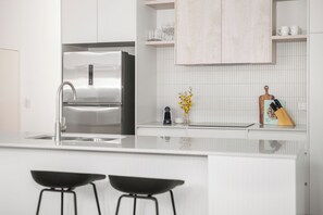 A modern kitchen with a stainless steel refrigerator, white cabinetry, a tiled backsplash, and a bar area with stools. - Foundry Lane Apartments by Urban Rest (Bowden)