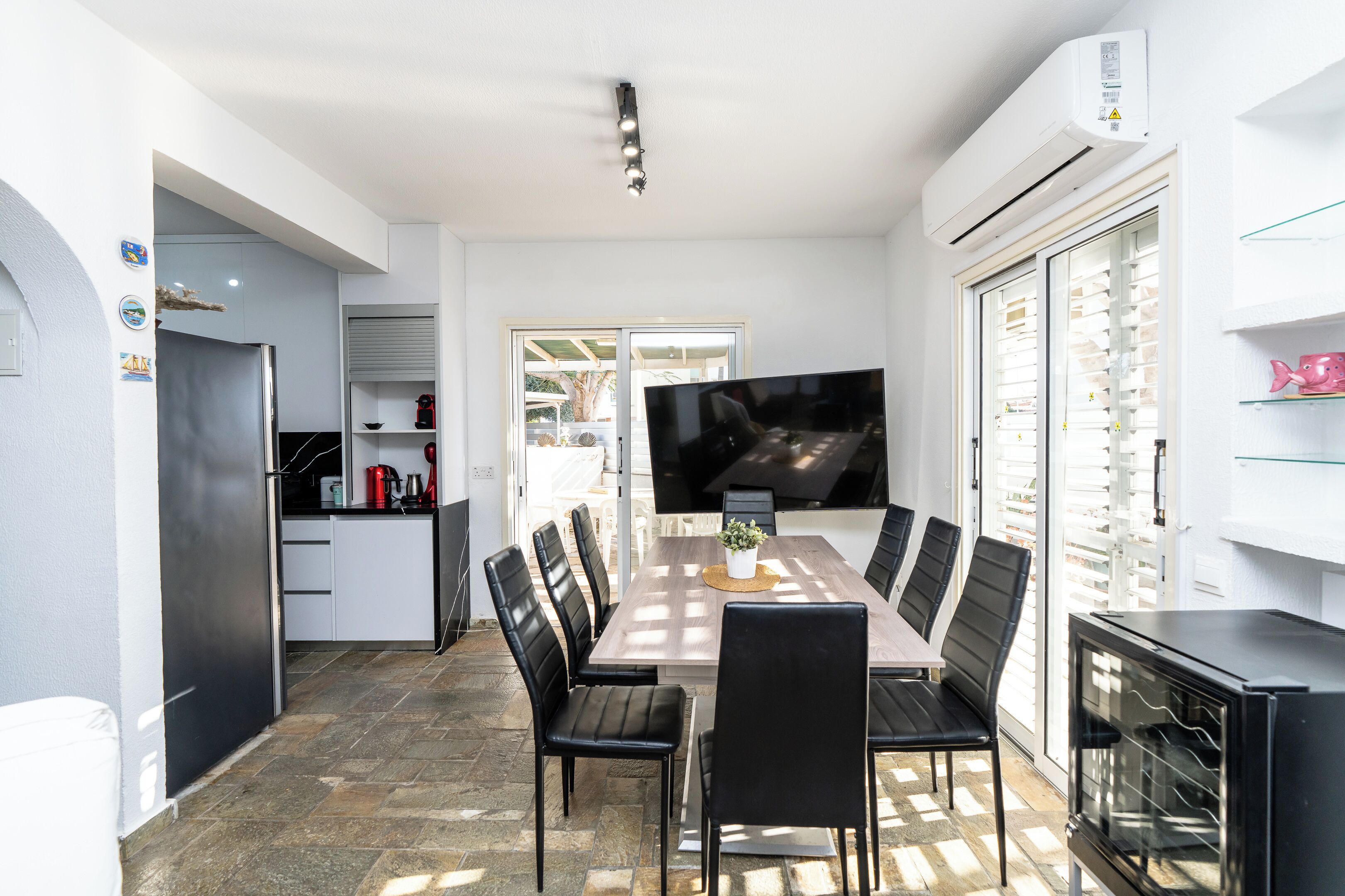 A modern kitchen with a dining area, featuring a large TV, a refrigerator, and a built-in oven.