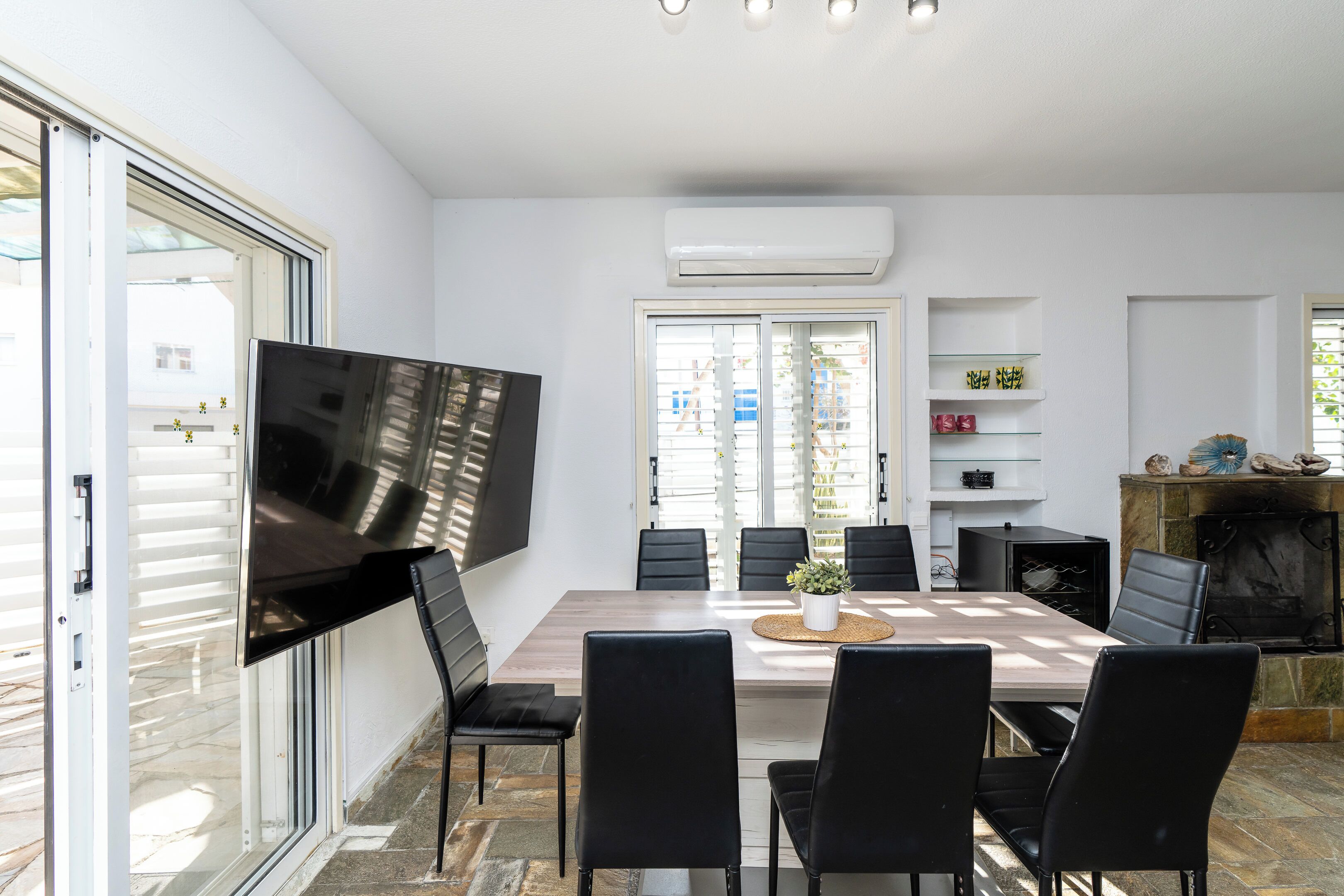 A modern dining area with a large table, black chairs, a flat-screen TV mounted on the wall, and a fireplace.