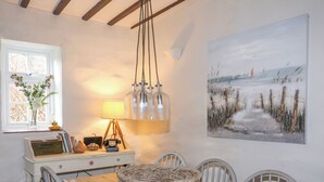 Een eethoek met een houten tafel, een rieten mand, witte stoelen, een lamp, een vintage radio en een schilderij van een strand.