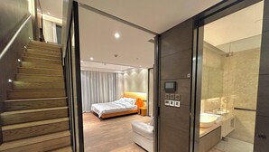 Une chambre d’hôtel moderne avec un escalier en bois, un grand lit, une table de chevet, un téléviseur intégré et une salle de bain dotée d’un grand miroir et d’un lavabo.
