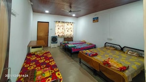A dormitory room with four single beds, a desk, a chair, a ceiling fan, and a window with curtains. - Alate's guest house (Bhor)