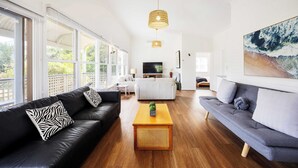 A spacious living room with a black leather sofa, a grey sofa, a wooden coffee table, and a large window providing a view of greenery.
